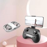 Drone de brinquedo branco com estrutura de proteção, controlado por um comando preto, exibido numa mesa rosa com um smartphone a mostrar uma corrida na praia.