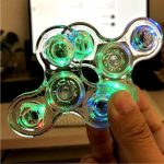 Fidget spinner transparente com LEDs verdes, azuis e vermelhos acesos, sendo segurado por uma mão, com fundo de computador e ambiente interno.