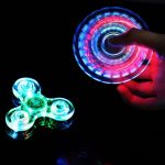 Fidget spinner luminoso com luzes LED coloridas em movimento, sendo segurado por uma mão, com fundo escuro.