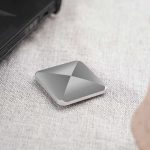 Dispositivo de armazenamento portátil com design quadrado e superfície metálica, colocado numa mesa de tecido claro, perto de um portátil preto.