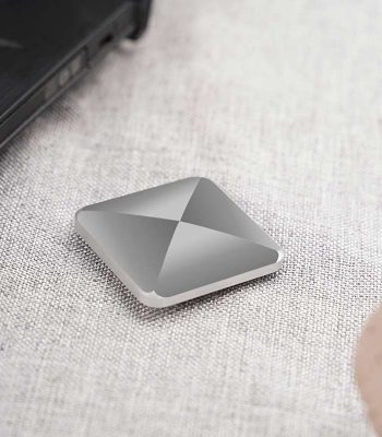 Dispositivo de armazenamento portátil com design quadrado e superfície metálica, colocado numa mesa de tecido claro, perto de um portátil preto.