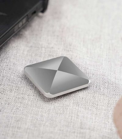 Dispositivo de armazenamento portátil com design quadrado e superfície metálica, colocado numa mesa de tecido claro, perto de um portátil preto.