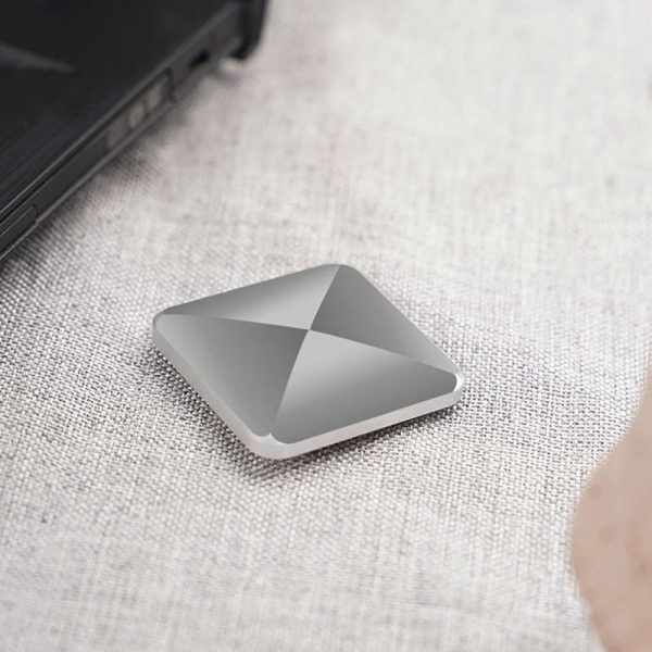 Dispositivo de armazenamento portátil com design quadrado e superfície metálica, colocado numa mesa de tecido claro, perto de um portátil preto.