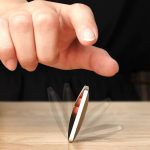 Disco de equilíbrio anti-stress com superfície metálica refletora, sendo equilibrado por uma mão, sobre uma mesa de madeira.