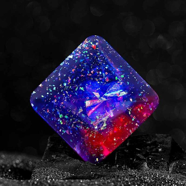 Cubo de vidro transparente com partículas brilhantes de várias cores, refletindo luzes azuis, vermelhas e verdes, sobre uma base escura.