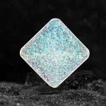 Cristal de vidro quadrado com efeito holográfico, refletindo cores iridescentes, colocado sobre uma superfície escura com fundo desfocado.