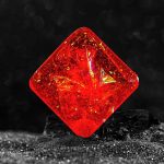 Cristal de vidro vermelho com reflexos dourados, em forma de cubo, colocado sobre uma superfície escura com fundo desfocado.