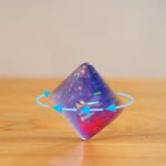 Dado de vidro com efeito holográfico, refletindo luzes coloridas, colocado sobre uma superfície de madeira, com setas azuis indicando rotação.
