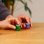 Duas mãos segurando brinquedos de ciência com efeito de plasma, um verde e um roxo, sobre uma mesa de madeira, com fundo desfocado de plantas.