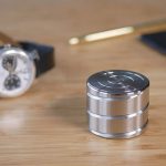 Giroscópio anti-stress de metal com superfície circular e linhas de design, colocado numa mesa de madeira com outros objetos ao fundo.