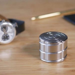 Giroscópio anti-stress de metal com superfície circular e linhas de design, colocado numa mesa de madeira com outros objetos ao fundo.