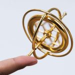 Giroscópio de precisão com estrutura metálica dourada, mostrando várias órbitas e eixos em diferentes ângulos, com uma pessoa segurando na base.