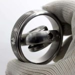 Giroscópio de metal com design circular e detalhes em linhas pretas e brancas, sendo segurado por uma mão com luva de tecido.