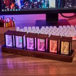 Relógio Nixie com seis tubos de vidro iluminados em cores diferentes, montado numa base de madeira, com fundo de computador e teclado.