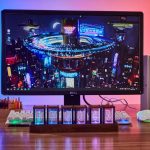Monitor de computador com fundo de cidade futurista e relógio Nixie em destaque na base, com teclado e caneta ao lado.