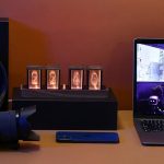 Quatro tubos Nixie com números visíveis, exibindo o tempo, numa base de madeira, ao lado de um portátil e um smartphone.