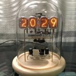 Relógio Nixie com tubos de néon exibindo o ano 2029, visível através de uma cúpula de vidro, com componentes eletrónicos internos e base de madeira.