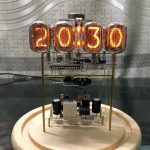 Relógio Nixie com quatro tubos de vidro exibindo a hora 20:13, com componentes eletrónicos visíveis e estrutura de suporte de metal, colocado sobre base de madeira.
