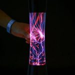 Tubo de vidro com plasma colorido em movimento, exibindo descargas elétricas azuis e rosas, com mãos segurando na parte inferior.