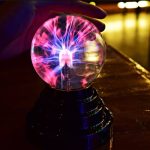 Lâmpada de plasma de 15 cm com relâmpagos coloridos dentro de uma esfera de vidro, iluminada por uma luz vibrante, colocada sobre uma base preta.