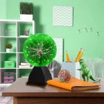 Lâmpada de plasma verde com relâmpagos visíveis dentro, colocada numa base preta numa mesa de estudo com objetos coloridos ao redor.