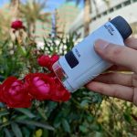 Microscópio portátil branco com lente de zoom, focando em flores vermelhas em jardim ao ar livre, com edifícios ao fundo.