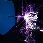 Descarga elétrica visível de uma mini bobina de Tesla com faíscas violetas e brancas, conectada a um dispositivo de controle, em ambiente escuro.