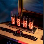 Relógio Nixie com quatro tubos de exibição de números em vidro, iluminados em tom laranja, colocado numa base preta sobre uma mesa de madeira.