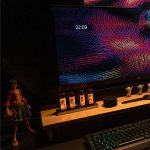 Monitor com efeito visual colorido e hora 22:09, acompanhado de objetos decorativos e teclado iluminado.