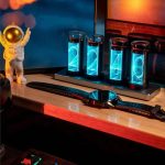 Relógio Nixie com visor de tubos de vidro iluminados em azul, acompanhado de uma figura de astronauta de brinquedo e uma pulseira de relógio no móvel de madeira.