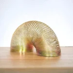 Mola Slinky dourada em forma de arco, colocada sobre uma superfície de madeira clara, com fundo branco.