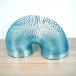 Mola Slinky de metal azul em forma de U, colocada sobre uma superfície de madeira clara, mostrando a sua estrutura espiral e flexível.