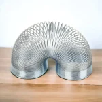 Mola Slinky metálica em forma de U vista de cima, com detalhes das espirais e acabamento brilhante, colocada sobre uma superfície de madeira clara.