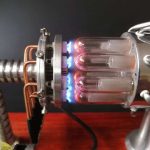 Reator de plasma com tubos de vidro transparentes, conectados a componentes metálicos e tubos de cobre, com faíscas azuis visíveis.