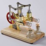 Modelo de motor Stirling com componentes de metal dourado, vidro e madeira, exibindo partes móveis e tubos de vidro com algodão, sobre base de madeira.