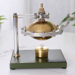 Motor Stirling com componentes metálicos dourados e prateados, base verde escura, mostrando o funcionamento com vapor de água.