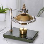Motor Stirling clássico com componentes metálicos dourados e prateados, montado sobre base verde, com uma pequena vela de teste abaixo, em ambiente de laboratório ou sala de aula.