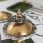 Detalhe do motor Stirling com componentes metálicos dourados e prateados, mostrando a câmara de combustão e o eixo de movimento.