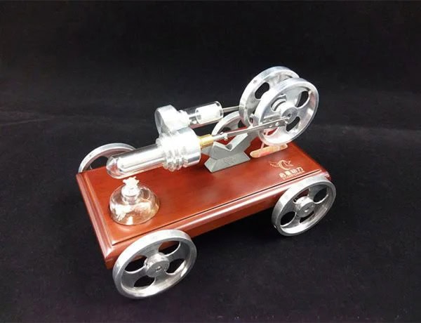 Modelo de motor Stirling em miniatura com rodas de metal, base de madeira e componentes de vidro e metal, exibido numa superfície escura.
