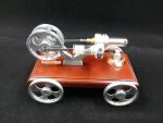 Modelo de motor Stirling com rodas metálicas, cilindro e sistema de pistão, colocado numa base de madeira.