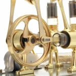 Motor Stirling com componentes dourados, incluindo um pistão, volantes e cilindros, usado em demonstrações científicas e projetos educativos.