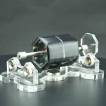 Motor Stirling solar com componentes de vidro e metal, apoiado em base de acrílico transparente, com foco na estrutura e detalhes do funcionamento.