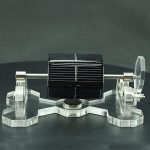 Motor Stirling solar com painel solar, suportado por estrutura de acrílico transparente, com componentes metálicos visíveis e detalhes de montagem.