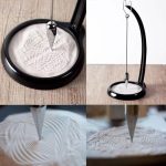 Pêndulo de areia de Foucault com base preta, mostrando o movimento da areia formando padrões no fundo branco, incluindo detalhes do pêndulo e da base.