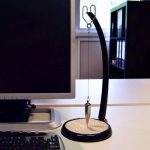 Pêndulo de Foucault com base preta, cabo curvo e esfera de metal pendurada, colocado numa mesa junto a um monitor de computador e uma estante de livros ao fundo.