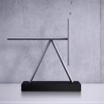 Pêndulo de Newton com bastão de metal, base preta e corda visível, em ambiente minimalista com parede de cimento ao fundo.