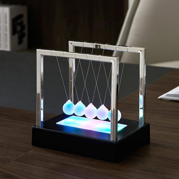Pêndulo de Newton com bolas brancas suspensas por fios finos, iluminado por luz LED azul na base, colocado numa mesa de escritório.
