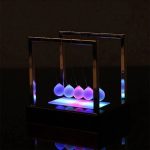 Pêndulo de Newton com bolas iluminadas por luz LED de cores diferentes, exibido em ambiente escuro.