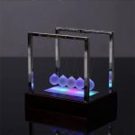 Pêndulo de Newton com bolas de vidro iluminadas por luz LED azul, exibido numa base escura com iluminação suave.