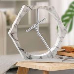 Relógio de mesa com estrutura de mármore branco e ponteiros metálicos, em estilo contemporâneo, colocado numa mesa de madeira com utensílios de cozinha ao fundo.